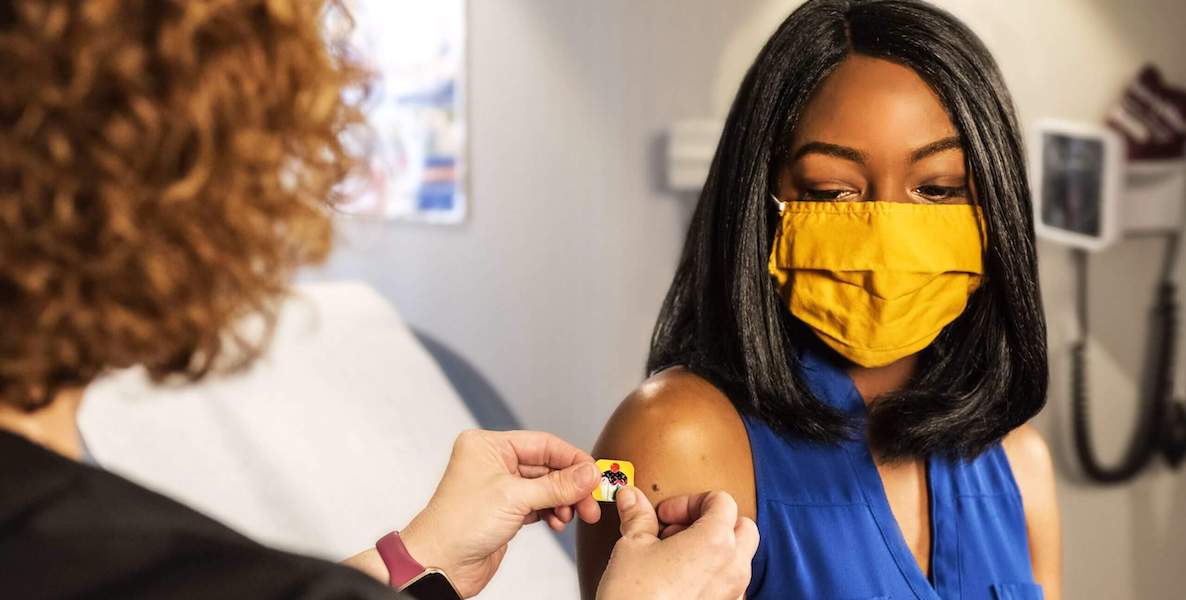 A Black woman wearing a yellow mask receives her Covid-19 vaccine from an nurse