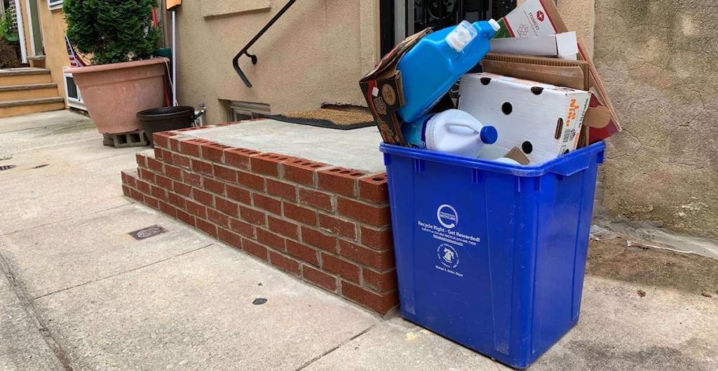 Blue recycling bin filled with recyclable items — detergent bottle, bleach bottle, cardboard — stands beside a rowhome stoop in Philadelphia.