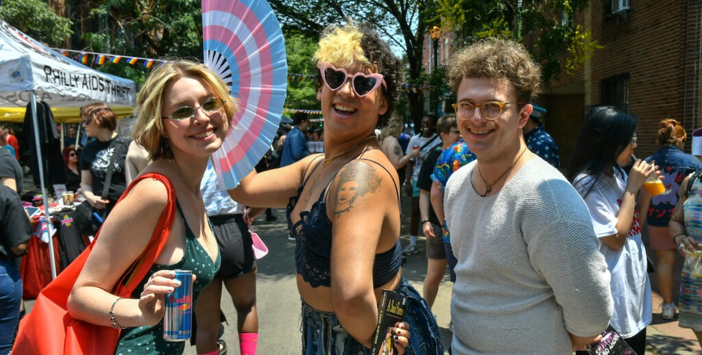 Three people stand outside, posing for the camera. One has short blonde hair and is wearing a V-neck green camisole. In the center is someone with short blonde and dark curly hair wearing sunglasses and holding a pink, blue and white fan. Last is a person with short curly brown hair wearing glasses and a long-sleeved white shirt. They are on Pine Street at Queerapalooza, an annual LGBTQIA+ Pride event put on by Philly AIDS Thrift at Giovanni's Room.