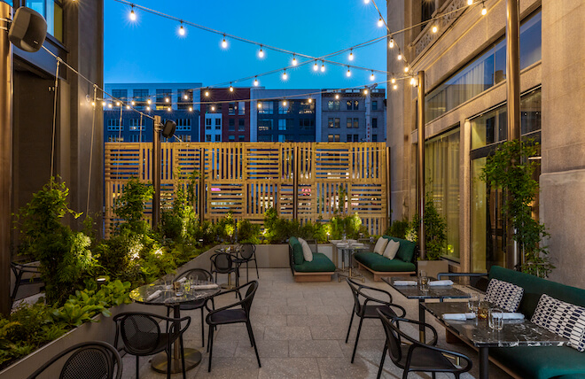 Black metal chairs and tables along with upholstered banquettes fill a small, planted courtyard with a wooden screen on one end and string lights overtop. This is the Wayward in Philadelphia.