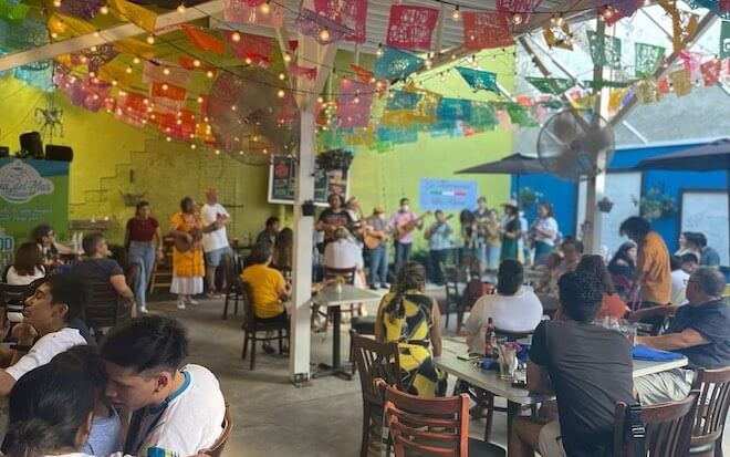 People sit at tables to watch a musical performance in the outdoor cafe of Alma del Mar, a Mexican-owned restaurant decorated with papel picado garlands.