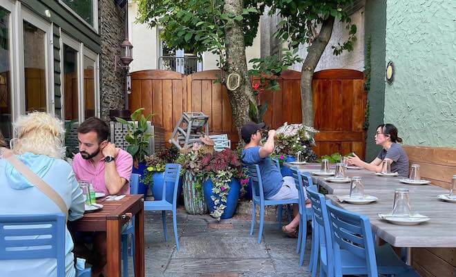 People sit outside in a courtyard with blue chairs and small tables. A tree stands in the background at Cantina Calaca Feliz in Fairmount, Philadelphia.