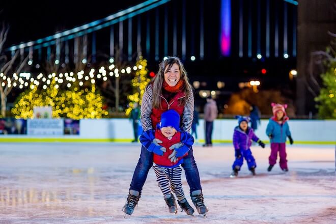 A smiling woman holds a young child while ice skating at the Blue Cross RiverRink. In the background is the Benjamin Franklin Bridge.