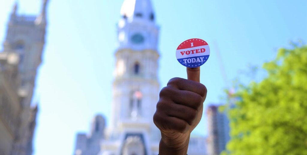 A brown hand poses in a thumbs up sign. At the top of the thumb is a red, white and blue "I Voted" sticker. Behind the hand in the distance are Philadelphia City Hall and a blue sky.