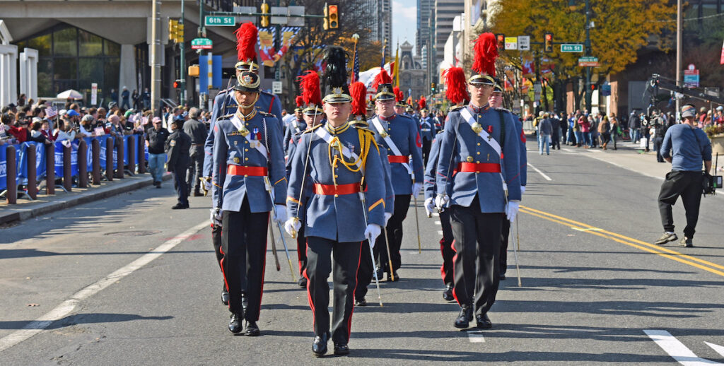 Uniformed marchers in blue coats, black pants and tall black hats march in the Veterans Day parade