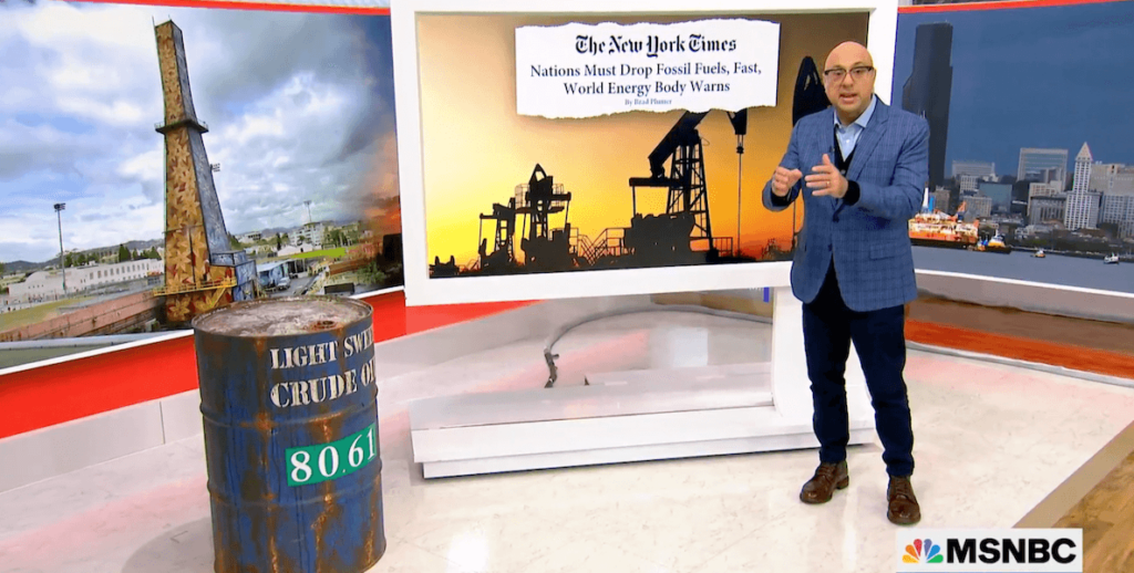 MSNBC host Ali Velshi stands before a screen featuring a New York Times article about the global rise of fossil fuel use. To his side stands an old oil barrel.