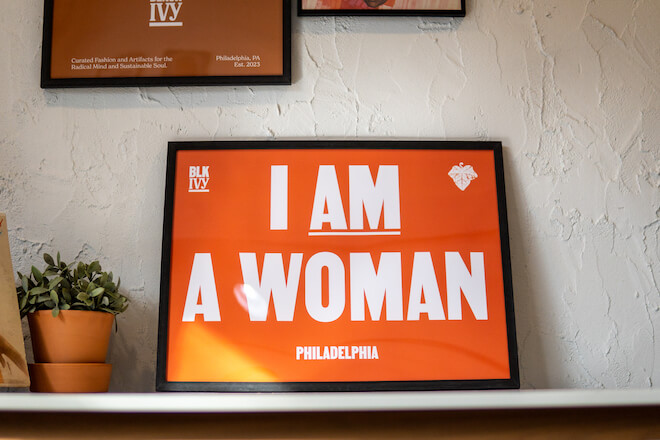 In the new vintage store Blk Ivy, an orange print in a black frame stands on a stable beside a plant. The print bears the words, "I AM A WOMAN." The letter "a" is underlined.