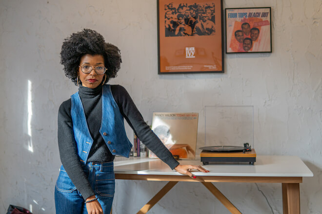 Kim McGlonn, wearing a denim vest, black turtleneck and denim pants, leans on a table against a white wall in her vintage boutique, Blk Ivy. On the table are a vintage album and record player. Hanging on the wall: A Blk Ivy sign featuring an old black-and-white photo and a Four Tops album, both framed.