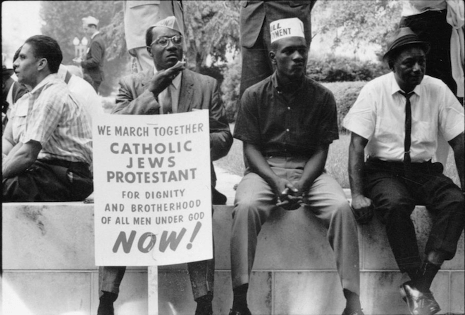 Photograph shows some participants in the civil rights march sitting on a wall resting, one holds a placard which reads, "We march together, Catholics, Jews, Protestant, for dignity and brotherhood of all men under God, Now!"