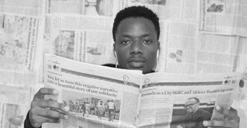 Against a backdrop of newspapers hung on a wall like wallpaper, a young Black man wearing earphones looks ahead while reading a newspaper that he is using both hands to hold open.