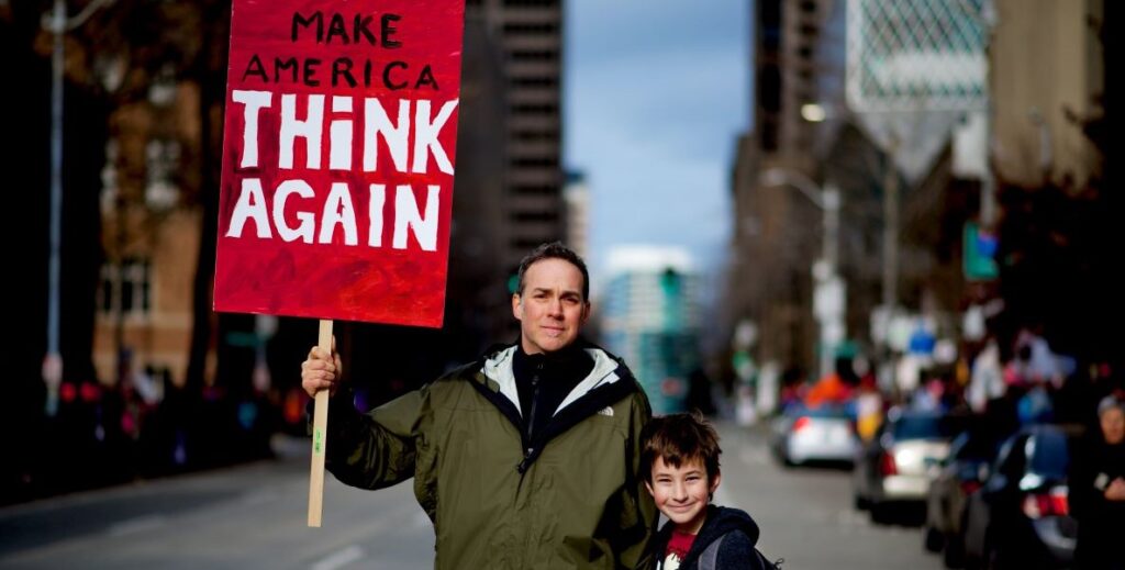 A man holds a "Make America Think Again" sign up as he stands beside a young boy in a street during a demonstration