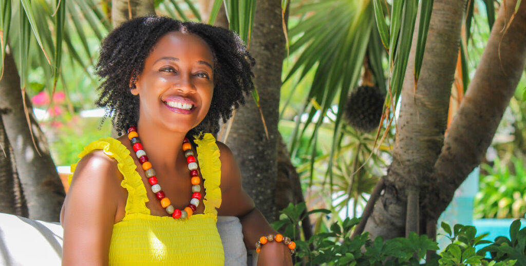 Mumbi Dunjwa of Naturaz smiles from an outdoor scene, wearing a necklace of large beads and bright yellow sundress. At her back: palm trees.
