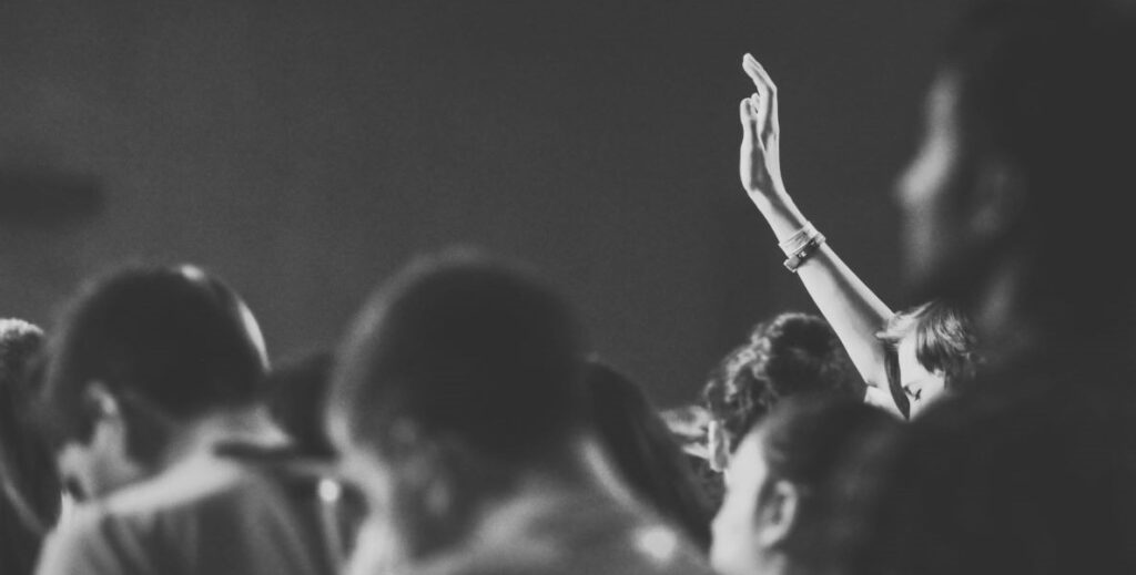 A black and white image of individuals in a class, from the neck up, with one hand raised in the background