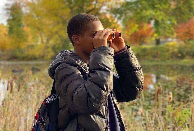 A young Black person with short hair and a black puffer coat and backpack holds binoculars to his face. He is standing in an autumn meadow.