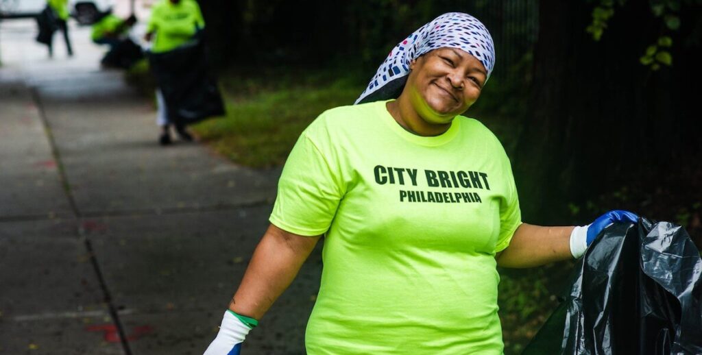 A woman wearing gloves and a bright green shirt that says "City Bright" holds a trash bag and smiles.
