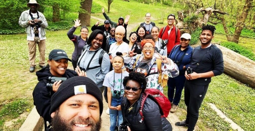 A group of mostly Black and Brown people with binoculars pose for a selfie outdoors.