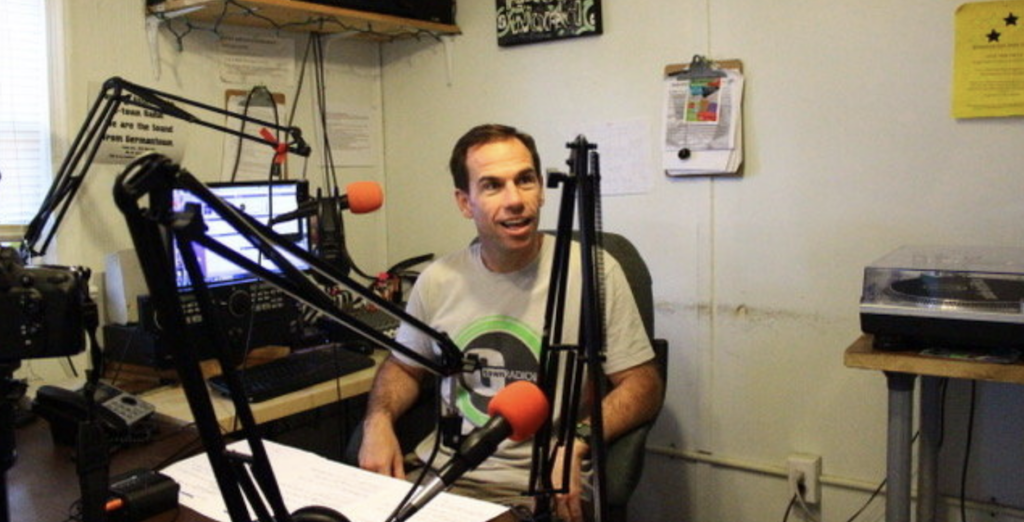 Photo of G-Town Radio founder Jim Bear, sitting and smiling behind microphones.