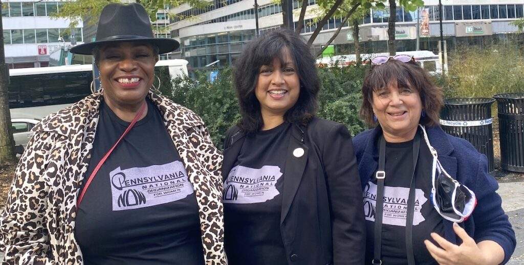 Three women stand together outside wearing matching Pennsylvania National Organization for Women t-shirts.