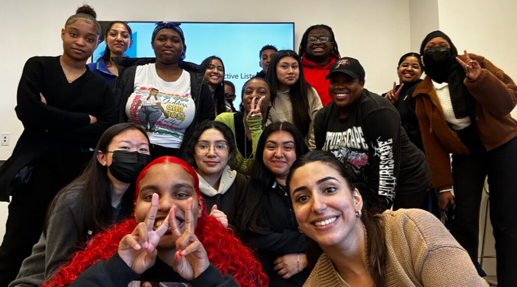 A group of smiling young people of color crouch, sit and stand. all facing forward.