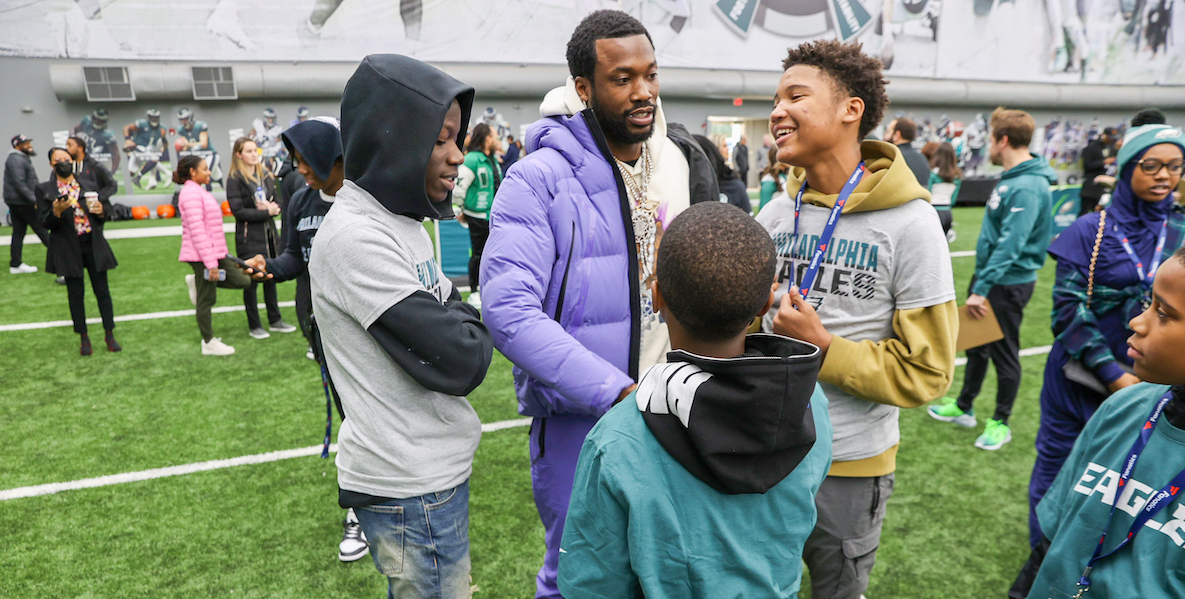 Meek Mills in a lavender puffer jacket, with children on the Eagles field.