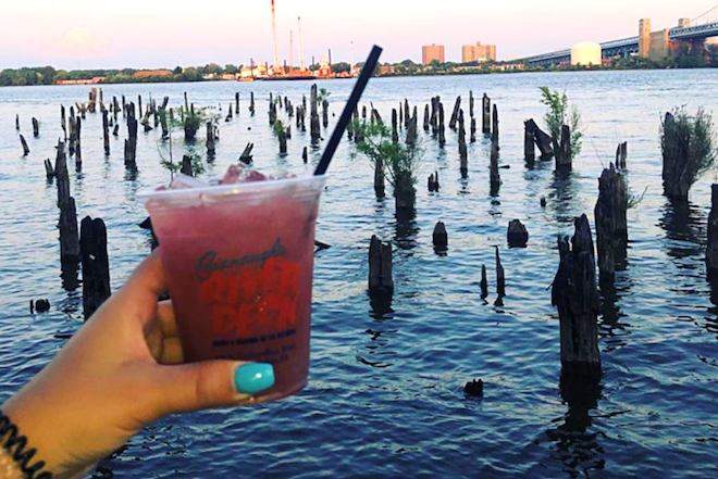 A hand with nails painted blue holds a red-colored drink in front of pilings on the Delaware River.