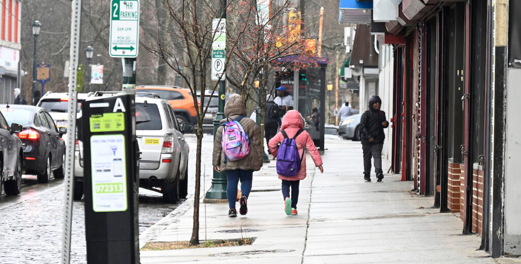 Two children wearing coats and backpacks walk along the sidewalk of Germantown Avenue.