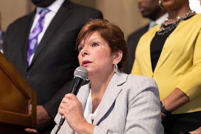 Seated, Pennsylvania Senator Christine Tartaglione from Northeast Philadelphia wears a beige suit jacket, has red-brown short hair, and speaks into a microphone. A group of people in suits stand behind her.