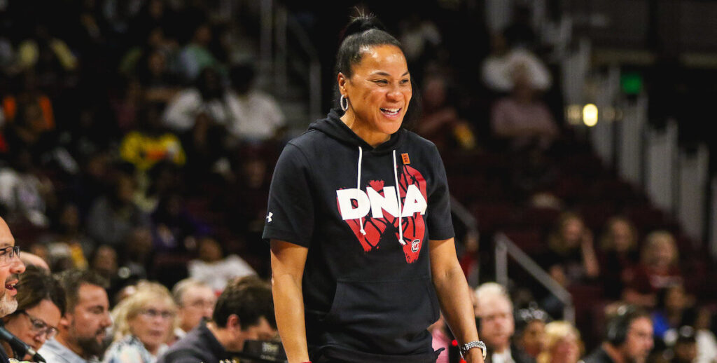 University of South Carolina basketball coach Dawn Staley, wearing a short-sleeved black DNA hoodie and her hair in a topknot, smiles from the sidelines.