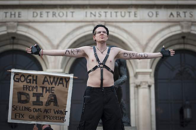 Jarod Bardon, a White male protestor, wears black belts over his bare torso while standing with his arms out in front of The Detroit Institute of Arts. His forearms bear the words "Art" and "Defend."