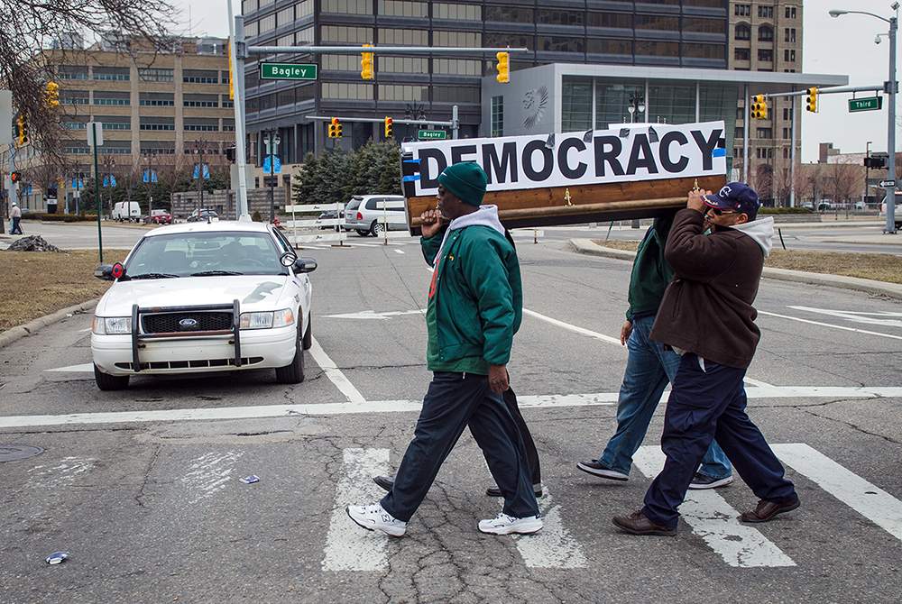 Four Black men carry a coffin bearing a sign that reads "DEMOCRACY" across a street in Detroit.