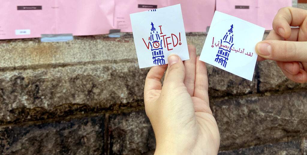 Two hands hold up "I Voted" stickers, backdropped by sample ballots.