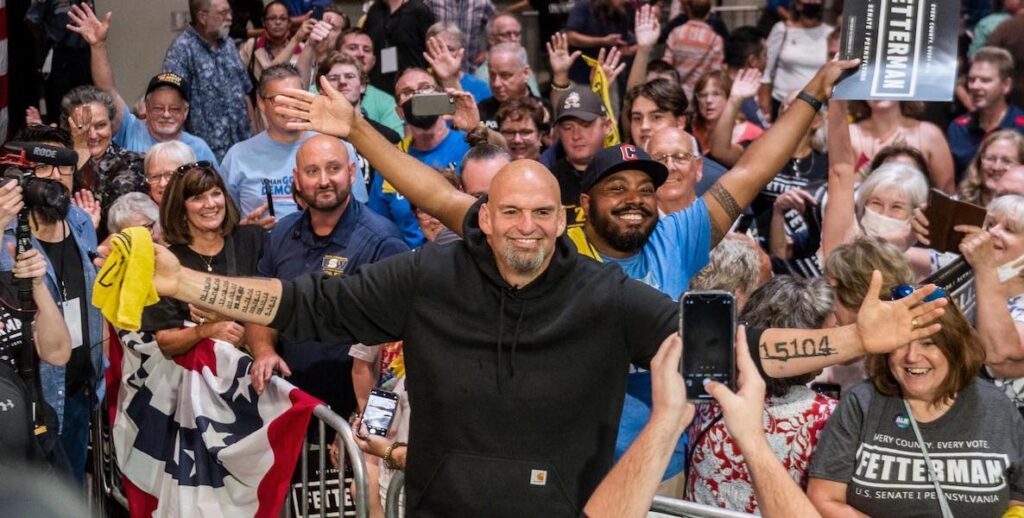 U.S. Senator John Fetterman, a tall, bald White man with wearing a black hoodie stretches out his tattooed forearms and smiles in front of a crowd cheering for him.