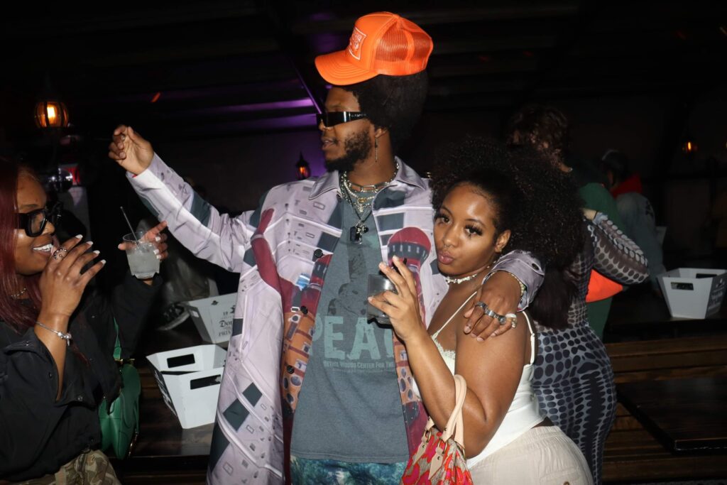 In a dark room, a couple stands shoulder to shoulder, arms around each other. To the left, the man wears a bright orange Mea Culpa trucker hat, dark sunglasses and colorful clothing. To the right, the woman in a cream tank and pants seems to be flashing the peace sign.