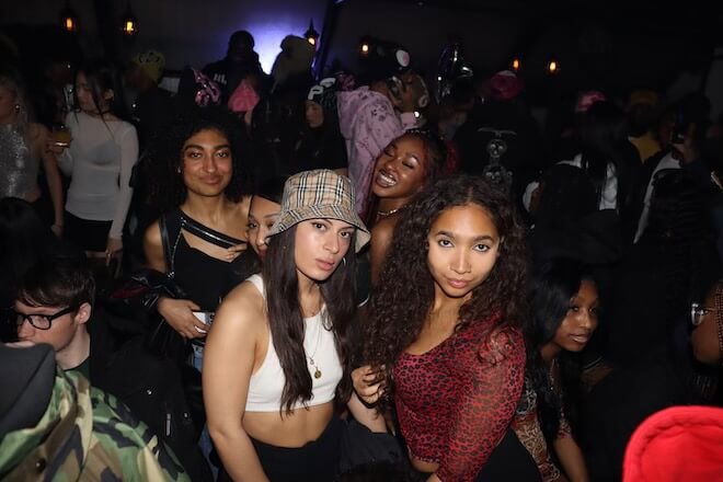 In a dark room with a purple light in back, a group of people dance. Before the camera, four women — one in a Burberry bucket hat — pose for the camera.