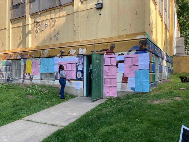 Pink and blue ballots are taped to an exterior wall of yellowed building with peeling paint. A woman stands by the ballots, looking at them. This is a polling place in the Cobbs Creek neighborhood of Philadelphia. 