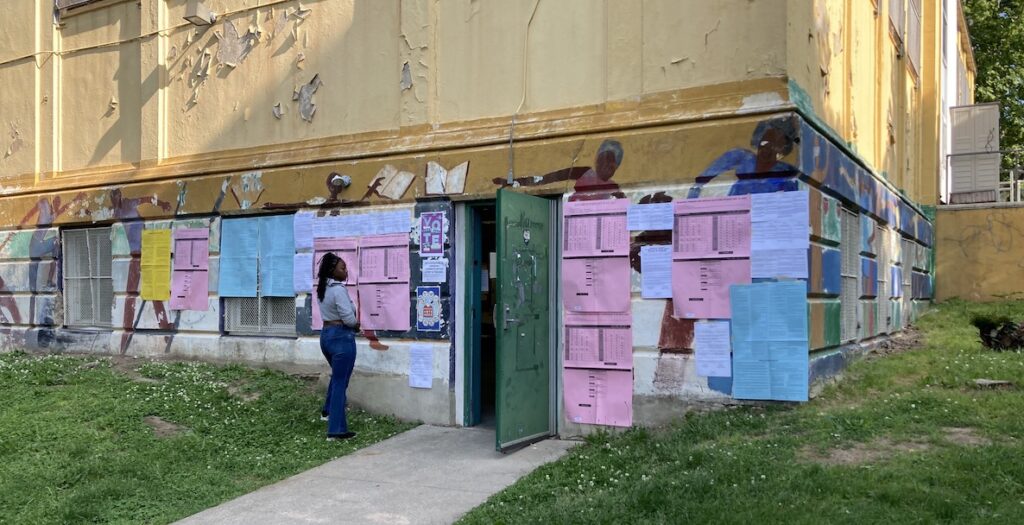 Pink, blue, white and yellow papers with voting information hang from a wall featuring a mural, part of a building with peeling yellow paint in the Cobbs Creek neighborhood of Philadelphia. A woman stands outside the building's open green door, facing the entrance.