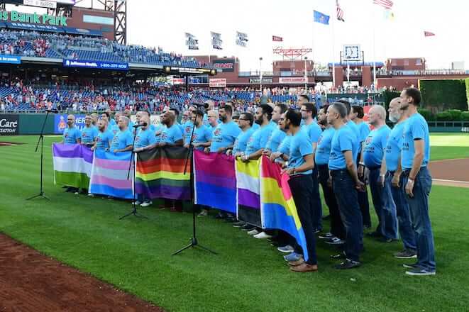 A chorus of men wearing blue t-shirts and jeans hold LGBTQIA+ flags on the field of Citizens Bank Park for Pride Night at the Philadelphia Phillies.