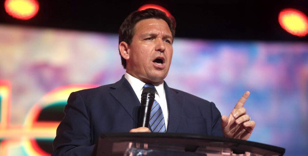 Florida Governor Ron DeSantis, wearing a navy blue suit and lighter blue tie, stands at a podium and speaks into a microphone at a 2022 rally.