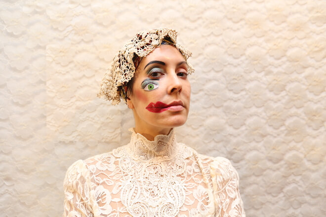 Artist Rose Luardo wears cream lace: a doily cap, and high-neck and puffed sleeves blouse. On one half of her face is a drawing of an eye and red lips.