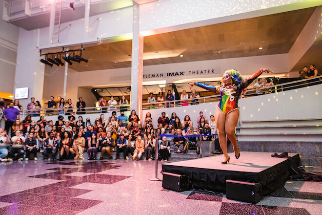 A drag queen wearing a rainbow bodysuit performs on a small stage before a crowd at The Franklin Institute.