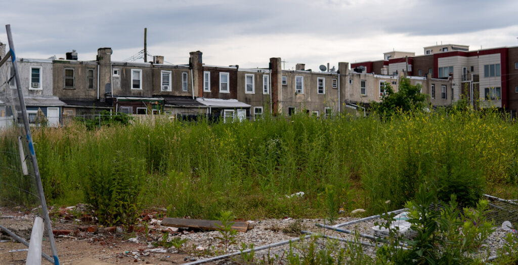 A chain-link fence has fallen onto a vacant lot where weeds grow tall. Behind the lot are rowhomes in Philadelphia.