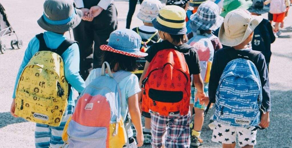 Four school children in summer hats and colorful backpacks walk to school in the morning sunlight