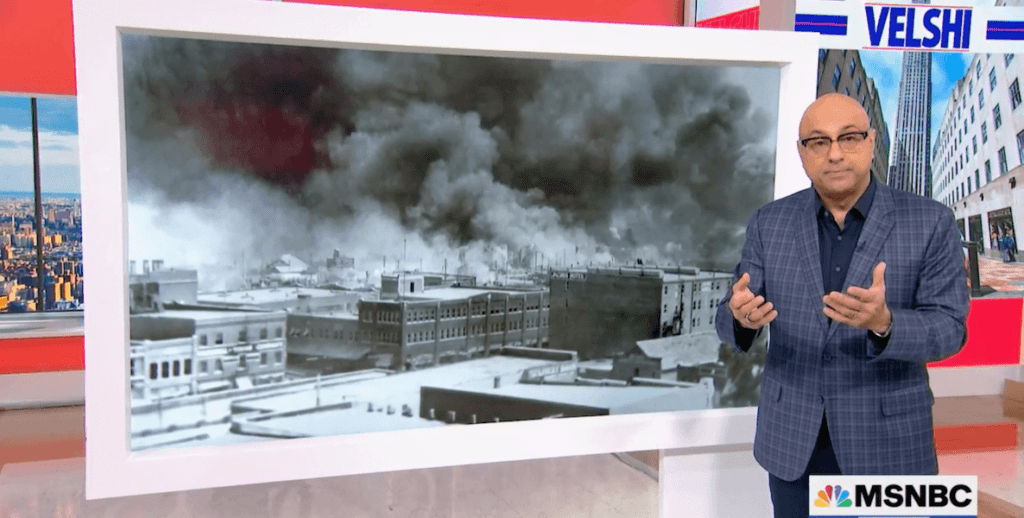 MSNBC host Ali Velshi stands before a black-and-white photo of a burning building to explain the Tulsa Race Massacre.