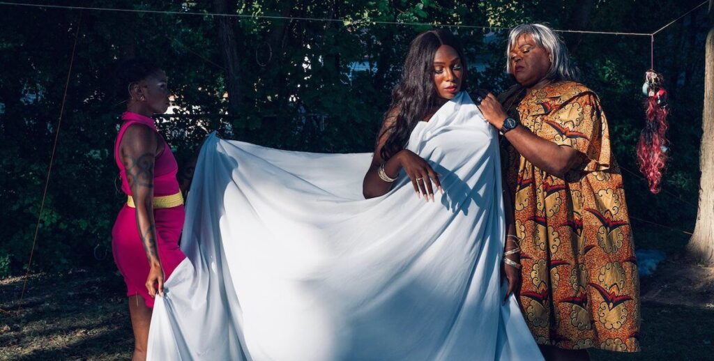 Two Black women in dresses stand on either side of a Black woman with long black hair. The two women appear to be holding two sides of the third woman's white sheet or dress.