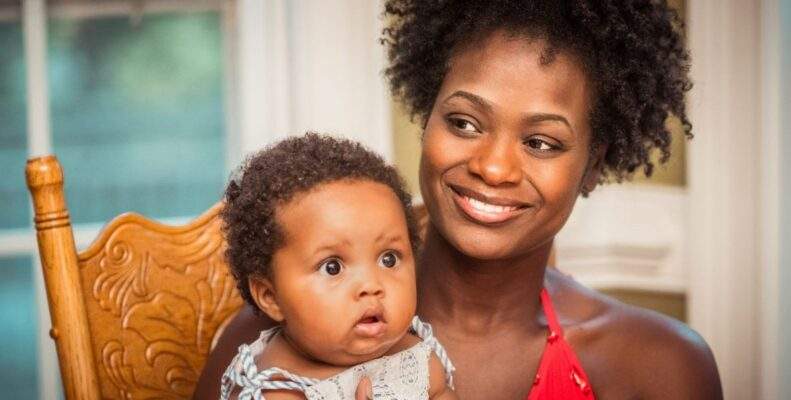 A Black mother smiles with her infant child on her lap staring adorably at something brightly colored, probably