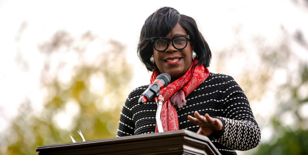 Cherelle Parker, a Black woman with mid-length hair wearing black-framed-glasses, a red scarf and black-and-white striped sweater, speaks behind a podium.