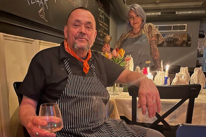 Chef Joncarl Lachman, wearing a striped apron and red banana around his neck, sits in a chair next to the chalkboard menu of his South Philadelphia restaurant, Dankbaar.