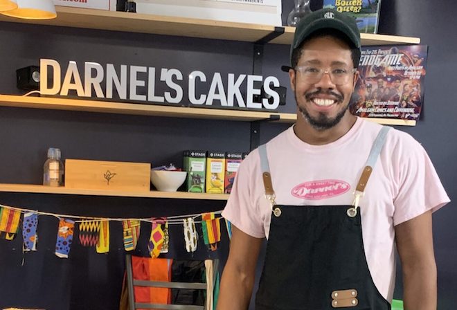 Kylie Cuffie-Scott, the co-owner of Darnel's Cakes, stands in front of a shelf bearing his bakery's name. He is a tall Black man wearing clear-framed glasses, a dark green baseball cap, pink Darnel's t-shirt, and black apron.