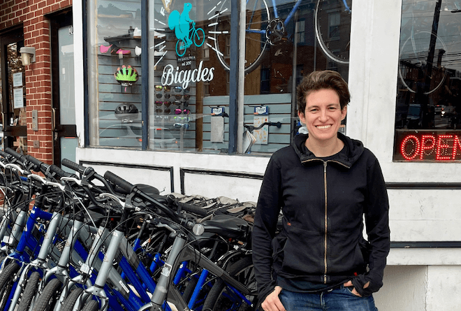 A smiling white woman with short black hair and wearing a black zipped-up hoodie stands in front of a row of bicycles parked on a sidewalk in front of a bicycle shop.