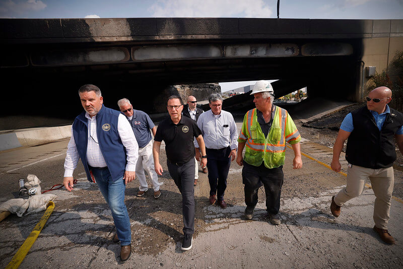PA Governor Josh Shapiro (center, wearing a black shirt, jeans and glasses) and his team walk from the site of the I-95 collapse.